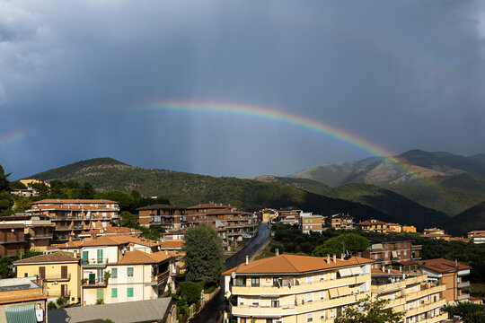 A bright full rainbow arcs over a sunlit town and green mountains under a dark stormy sky. Dramatic weather contrasts inspire deep feelings of hope, peaceful community renewal and natural harmony.