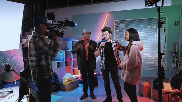 A young reporter interviews movie stars inside a modern film studio with a professional crew filming the interaction amid set equipment and bright lighting.