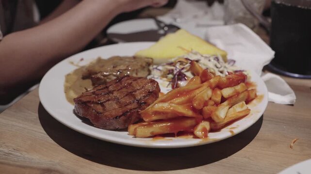 A steak being trimmed of its burnt parts with french fries, ketchup, coleslaw, and toast. Close-up of a person cutting the juicy meat with a knife and fork at a restaurant dining table.