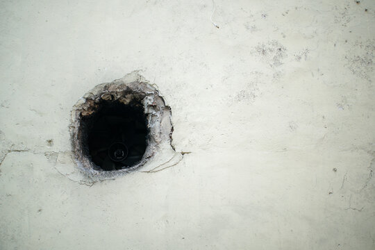 Close up of a dark circular hole in an old concrete wall with cracks and damaged plaster, representing building deterioration, construction defects, repair, renovation, maintenance.