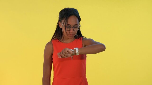 A young black woman wearing an orange sleeveless dress and eyeglasses checks the time on her wristwatch in a studio with a solid yellow background.