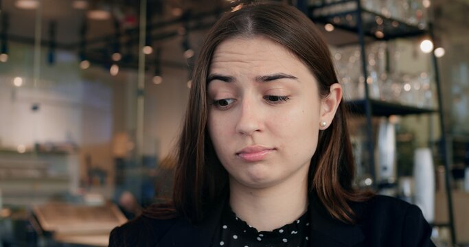 Brunette woman shows doubtful face during quiet moment. Contemplative young adult furrows brow thoughtfully, glancing downward with skeptical demeanor and visible uncertainty inside modern space.