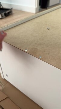 Man hammering two nails in a row along the edge of a white cabinet back panel, vertical close-up. Vertical close-up of hands driving two nails into the fiberboard back of a cabinet.