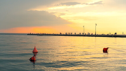 Serene sea view at sunset with a long pier, silhouettes of people, and floating orange buoys © NORMA