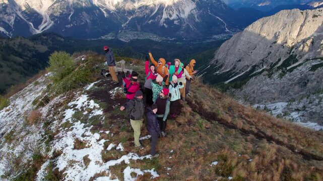Drone view shows cheerful group of hikers posing and waving on mountain summit, surrounded by dramatic rocky peaks, alpine valleys and patches of snow in Dolomites, Italy