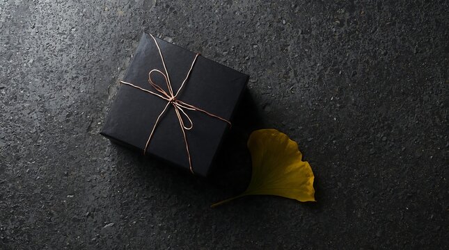 Elegant minimalist black gift box tied with a thin copper wire and a simple bow, placed on a dark wet textured stone surface next to a single yellow ginkgo leaf for a luxury seasonal aesthetic.