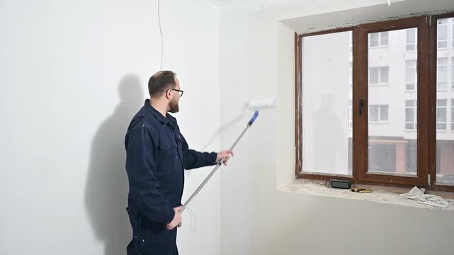 Man paints ceiling with roller extension pole inside modern apartment. Interior decorating, wall finishing, and residential repair work in progress