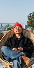 Obraz premium Man relaxing on rooftop wicker chair wearing a red beanie with city skyline background for lifestyle banner and relaxation design