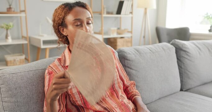 Woman cooling herself with fan in heat. Sitting on sofa at home, she seeks relief from hot indoor temperature and overheating during summer. Everyday indoor cooling discomfort concept.