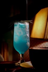 Refreshing Blue Lagoon Cocktail with Ice Cubes and Rosemary Garnish in Glass on Warm Bar Atmosphere © Rara