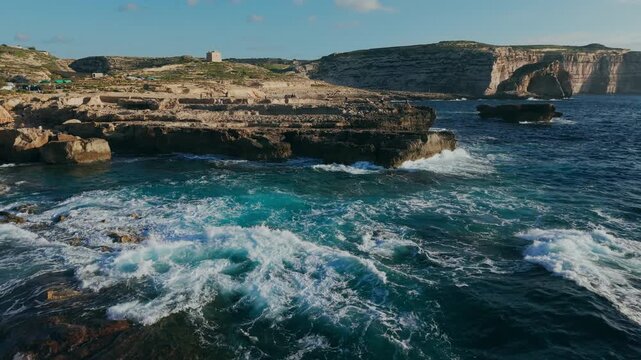 Aerial view of turquoise waves swirling over the rocky shore at Dwejra, Gozo, Malta. White foam, sea cliffs, Fungus Rock and watchtower in the background.