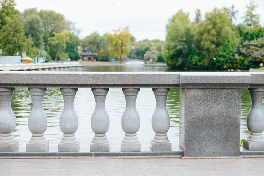 A stone railing made of balusters encloses a walkway next to a calm lake. Lush green trees and small boats are visible in the background, creating a natural setting