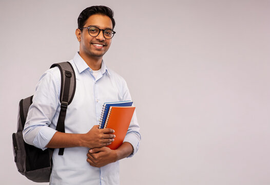 Smiling Indian male university student holding colorful notebooks and wearing backpack on light gray studio background