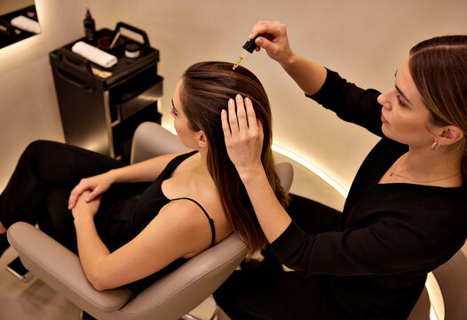 Hairstylist Applying Hair Serum Scalp Treatment to Client in Modern Salon Warm Interior Lighting