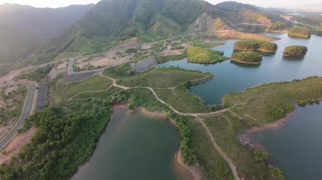 Drone glides forward over a still lake dotted with small green islands as a winding road curves along forested mountain slopes in soft warm light.