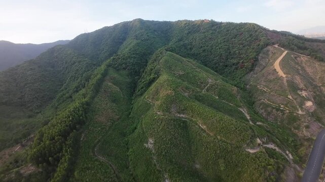 Aerial drone footage moves over lush mountain slopes with a winding paved road and exposed earth in warm evening light.