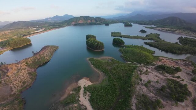 Aerial drone footage glides over a broad mountain reservoir with forested islands, layered green hills and cloudy evening skies.
