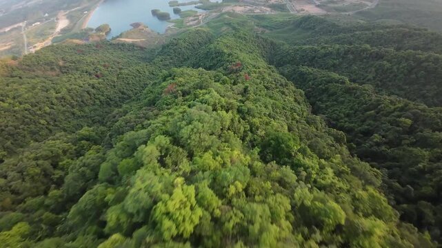 Aerial drone footage flies over dense forested mountain ridges toward a reservoir with landscaped islands and winding roads in warm evening light.
