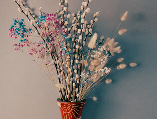 A vibrant bouquet of dried flowers, including pussy willow branches, pink and blue baby's breath, and fluffy bunny tail grass, elegantly fills a patterned ceramic vase against a muted grey background. © Kos6ka