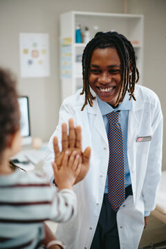 Vertical shot of happy African American young adult pediatrician giving high five to little child during medical checkup. Concept representing pediatric healthcare and clinic services