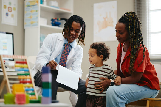 African American male pediatrician writing medical notes during checkup consultation with African American toddler boy and mother. Suitable for pediatric healthcare marketing
