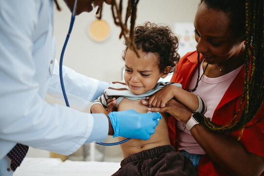 African American pediatrician examining toddler child chest with stethoscope beside mother during clinic visit. Image suitable for pediatric healthcare campaign and family medicine advertising