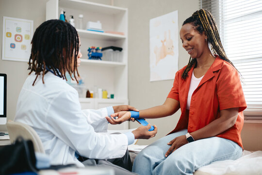 African American doctor wrapping bandage on wrist of smiling African American woman in clinic. Visual represents healthcare, suitable for medical clinic marketing