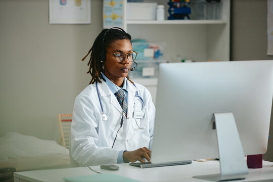 African American young adult male pediatrician wearing headset working at computer in clinic. Useful for telemedicine services, pediatric healthcare software, and online medical consultation