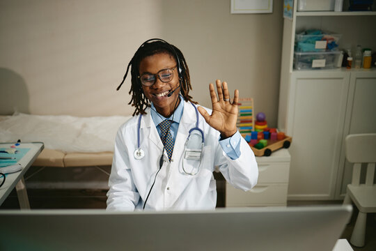 Young adult African American male pediatrician with headset waving hand during online telemedicine video call on computer. Useful for digital pediatric telehealth consultation services