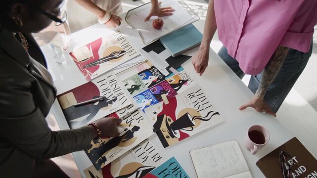 Diverse creative team reviewing and arranging printed book cover designs on table during brainstorming meeting. Useful for design agency, publishing, and creative teamwork campaigns