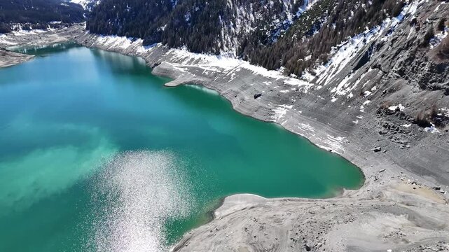 steep drawdown bank curves around turquoise lai da marmorera cove snow patches cling forested slopes above exposed gravel shoreline glacial reservoir water aerial drone footage graubunden switzerland