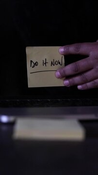 A vertical, slow-motion shot of a person's hands as they stick a sticky note with the message &ldquo;Do it now&rdquo; on a computer screen 