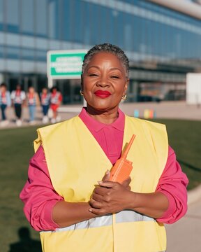 Black woman fire warden in a yellow hi-vis tabard holding a radio, standing calm at a sunny assembly point for workplace safety and EHS software