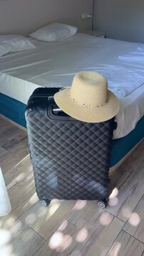 Black suitcase with a straw hat placed on top, positioned next to an unmade bed with white sheets in a bright, sunlit hotel room interior