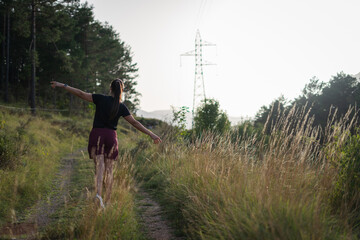 Fototapeta premium Young woman walking with arms outstretched on a rural path near power lines
