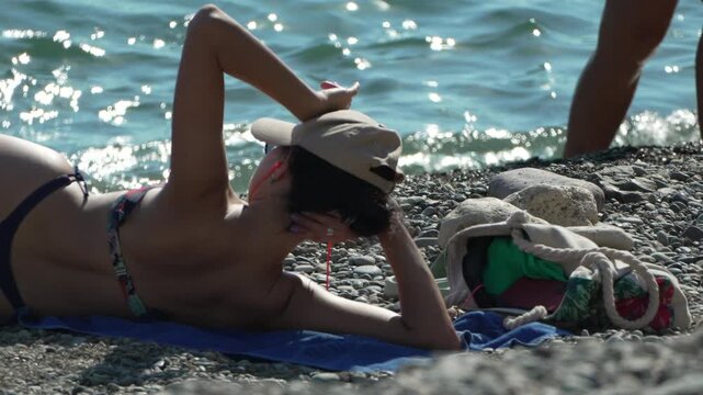 Sunbathing woman wearing baseball cap relaxing on pebbled shore by sparkling ocean waves. Close up steady shot.
