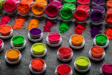 Obraz premium Vibrant colored powders lined up in small containers seen from above on a dark surface. Bright colors are spread across the dark surface with various containers filled with powders.