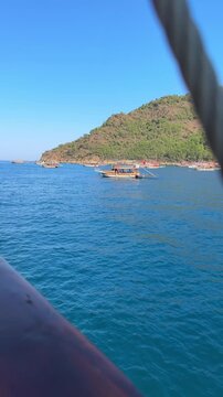 Scenic view of boats navigating turquoise waters near a lush green hillside, with a close-up of wooden railing and yellow ropes on a sailing vessel