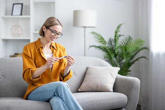 Pregnancy test result, happy woman checking medical device at home, family planning and fertility concept, smiling female sitting on a living room sofa, positive news and motherhood anticipation.