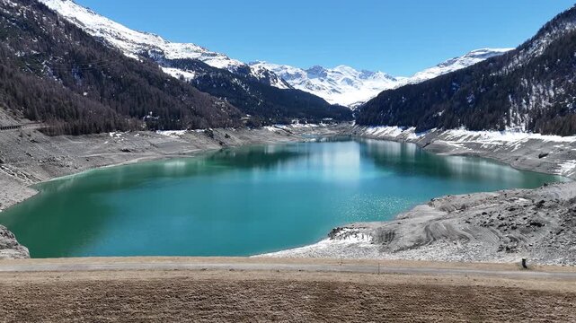 cyclist crosses dam crest road overlooking turquoise lai da marmorera reservoir exposed drawdown shoreline snowcapped peaks tower valley handheld footage graubunden switzerland julier pass