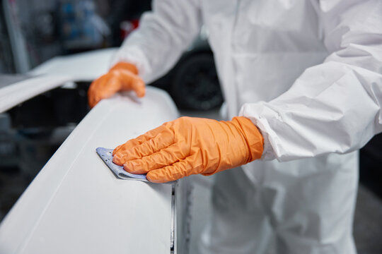 Automotive technician sanding car body panel with orange gloves and protective coverall