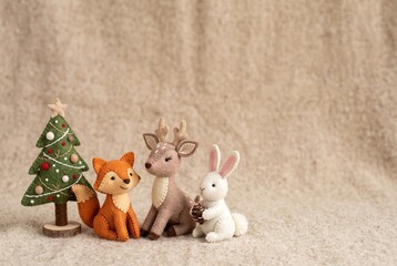 Fototapeta premium Felt figures of a fox, deer, and rabbit gather around a small Christmas tree. They sit on a neutral-colored background. This scene captures a holiday theme with copy space