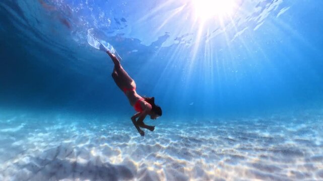 Underwater freediving adventure featuring a woman in a red bikini exploring a sandy ocean floor. Wide tracking shot.