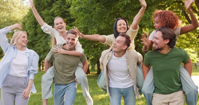 Friends piggyback in park, laughing, playful couples. Cheerful group of people outdoors shares fun and captures togetherness, leisure and team spirit. Joyful friendship and bonding concept.
