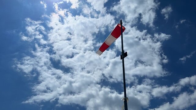 Red and white windsock flutters against a bright blue sky filled with fluffy clouds, showcasing the wind direction and weather conditions in an outdoor setting