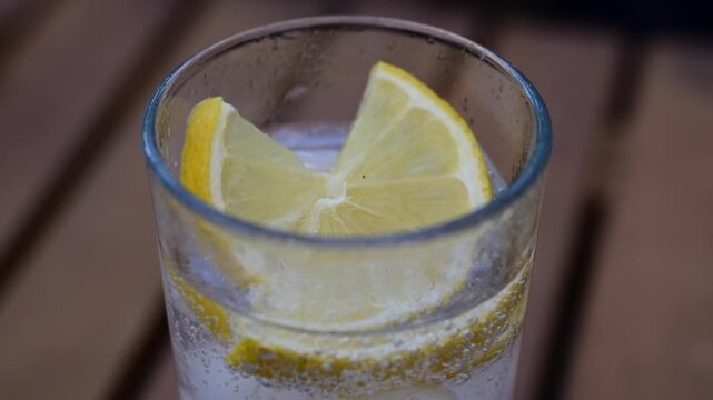 Fresh, juicy lemon slices rest inside a glass of bubbly, sparkling water effervescing on an outdoor wooden table.