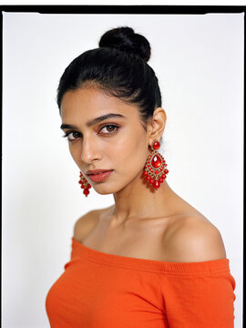 South Asian woman with bun hairstyle wearing orange off shoulder top and red chandelier earrings on white background