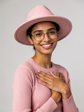 Smiling Indian woman wearing pink fedora hat and glasses holding hands over heart in sincere gesture studio