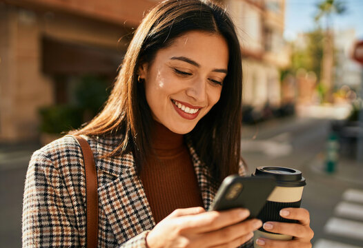 Smiling young woman using smartphone and holding coffee cup in sunny urban city street while commuting outdoors
