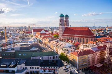 A panoramic view of Munich featuring the iconic Frauenkirche with its twin green-domed towers at sunset, surrounded by historic rooftops and the cityscape under a bright blue sky. © IRStone
